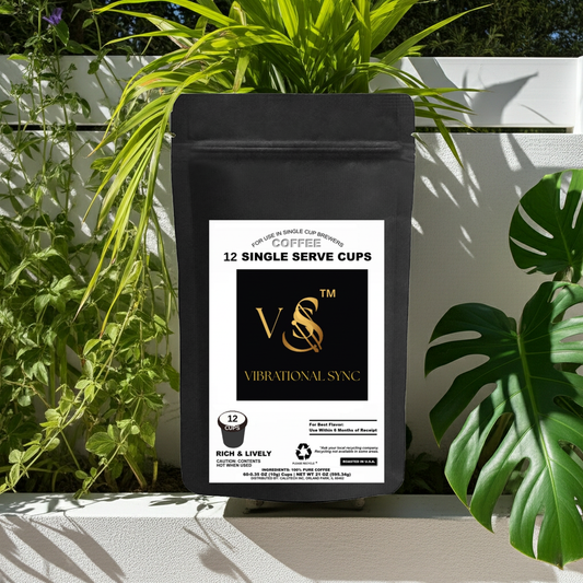 A black stand-up coffee bag labeled VVS Vibrational Sync, 12 single-serve cups, placed outdoors on a white surface with green plants and leafy branches framing the package.